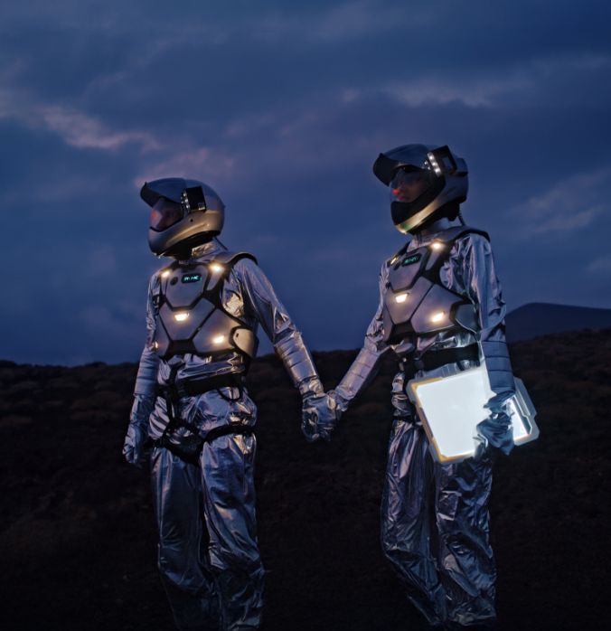 Two people in silver space suits hold hands